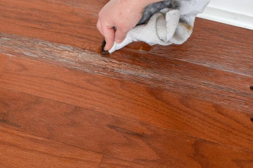 A hand is wiping liquid from a damaged hardwood floor surface with visible finish wear and moisture marks.