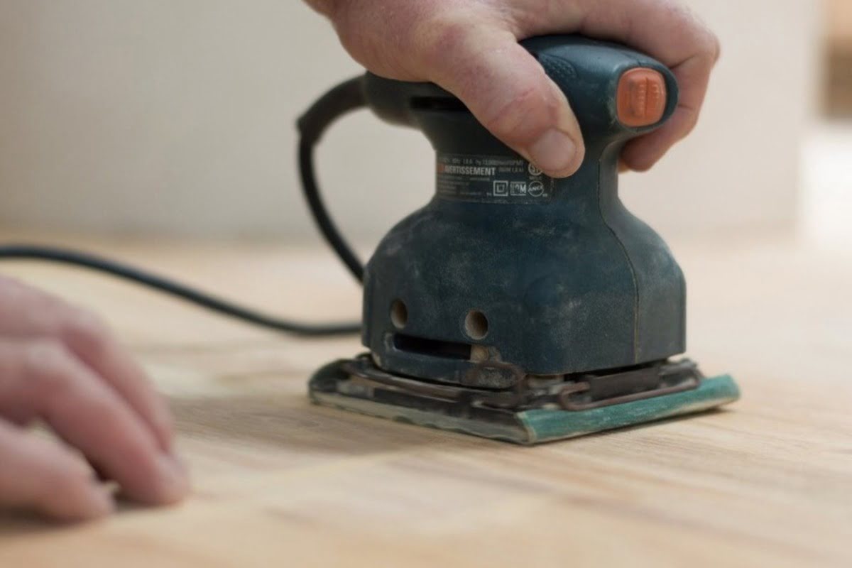 A handheld power sander is smoothing the surface of a hardwood floor during the refinishing process.