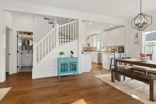An open concept home interior features dark hardwood flooring, white walls, a staircase, and a bright kitchen and dining area.