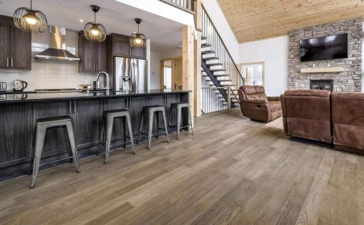 An open concept interior features wide plank wood flooring, a dark kitchen island, a staircase, and a living area with a stone fireplace.