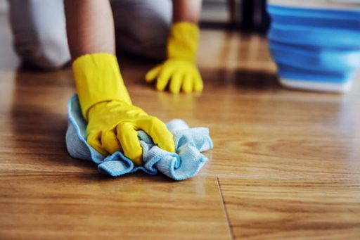 A person wearing yellow gloves is cleaning a hardwood floor with a cloth to prepare the surface before floor repair or refinishing.